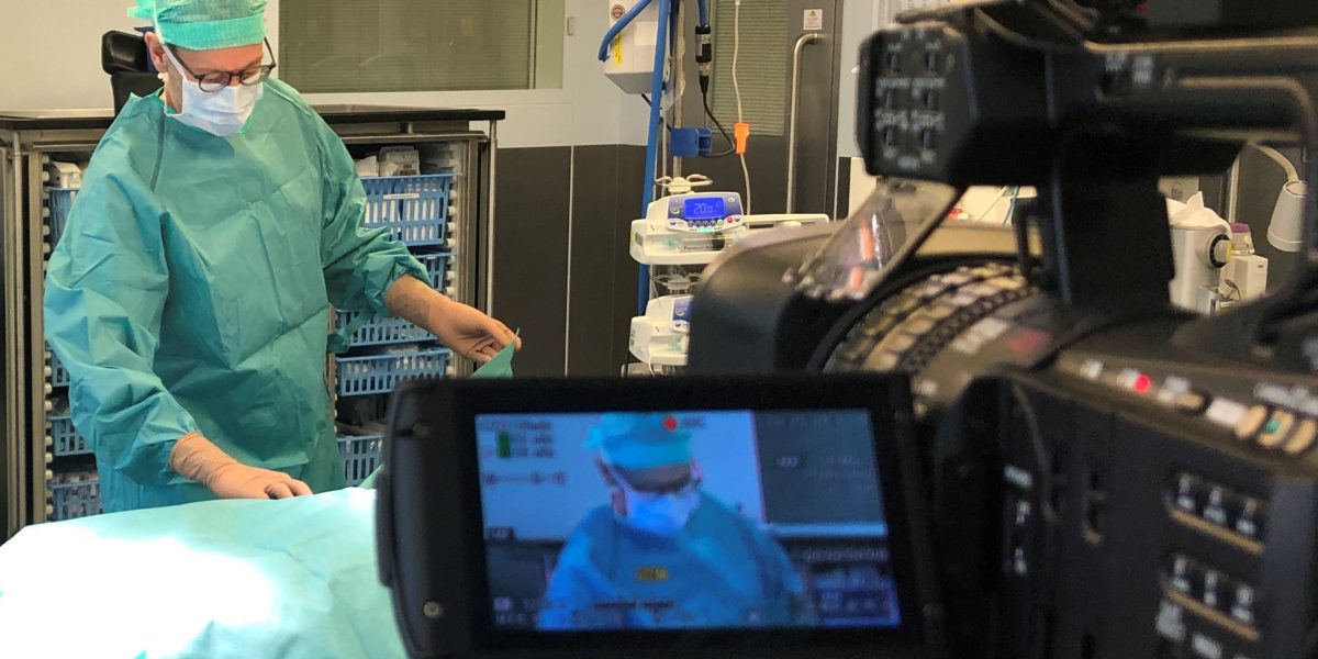 Chirurg in OP-Kleidung wird während einer Operation von einer Kamera gefilmt. Monitor zeigt Aufnahme.