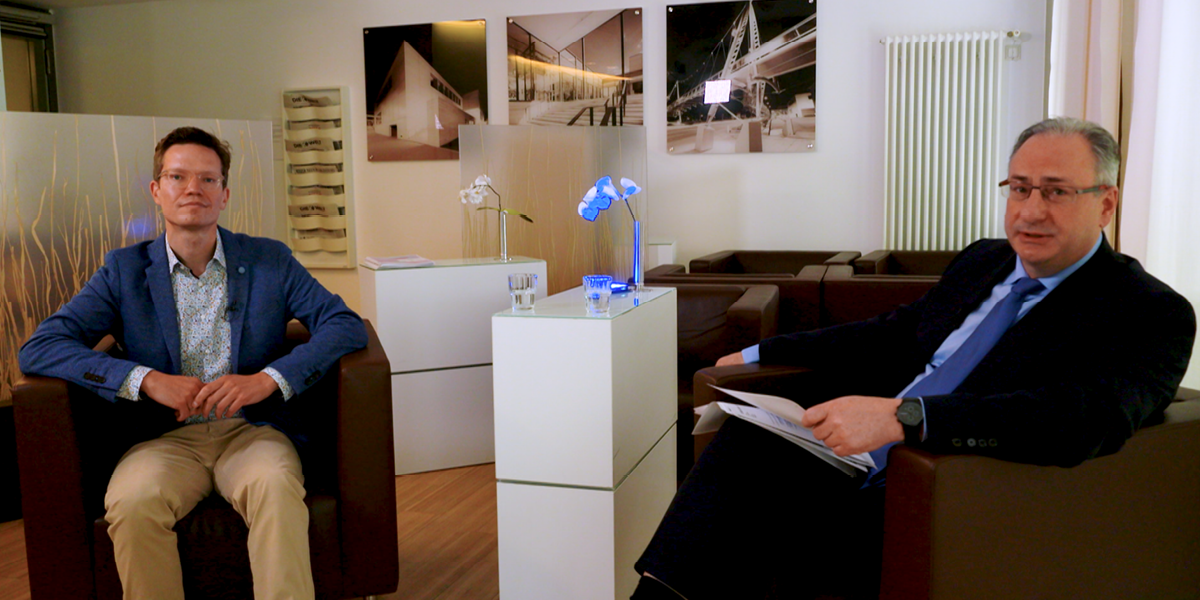 Zwei Männer in Anzügen sitzen in einem modernen Büro mit Fotos an der Wand, flankiert von weißen Möbeln und Orchideen.