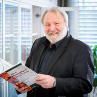 Älterer Mann mit Bart in Anzug hält ein medizinisches Buch. Er steht vor einem Regal und schaut lächelnd in die Kamera.