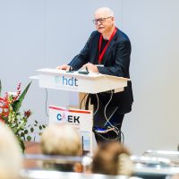 Ein Mann hält eine Rede am Rednerpult bei einer Konferenz, Blumenarrangement auf der linken Seite.
