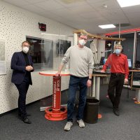 Drei Männer mit Masken in einem Radiostudio von Radio Essen, stehend neben Tischen und Mikrofonen.