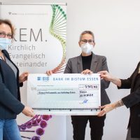 Drei Personen mit Masken überreichen in Essen einen symbolischen Scheck über 9.913,47 Euro vor einem Banner.