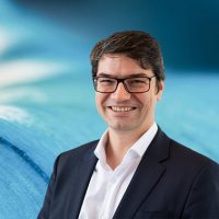 Ein lächelnder Mann mit Brille in Anzug vor unscharfem, blauem Hintergrund.