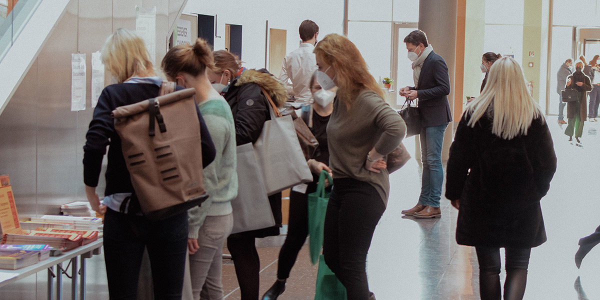 Personen mit Masken in einer Halle, einige schauen auf einen Tisch mit Büchern und Informationsmaterialien.