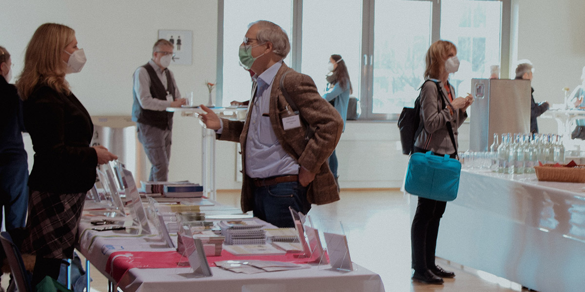 Personen mit Masken bei einer Konferenz; Bücher auf einem Tisch und Getränke auf einem anderen.