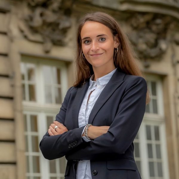 Eine Frau in einem dunklen Blazer steht mit verschränkten Armen vor einem Gebäude mit großen Fenstern.