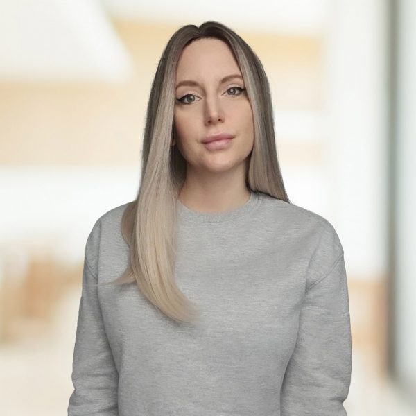 Eine Frau mit langen, glatten Haaren und grauem Pullover steht vor einem unscharfen, hellen Hintergrund.