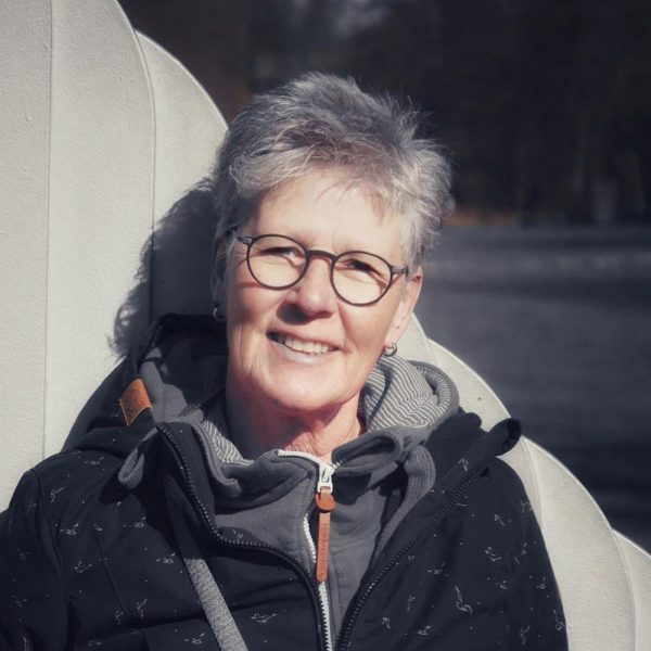 Ältere Frau mit Brille lächelt in die Kamera, sitzt im Freien, trägt eine warme Jacke.