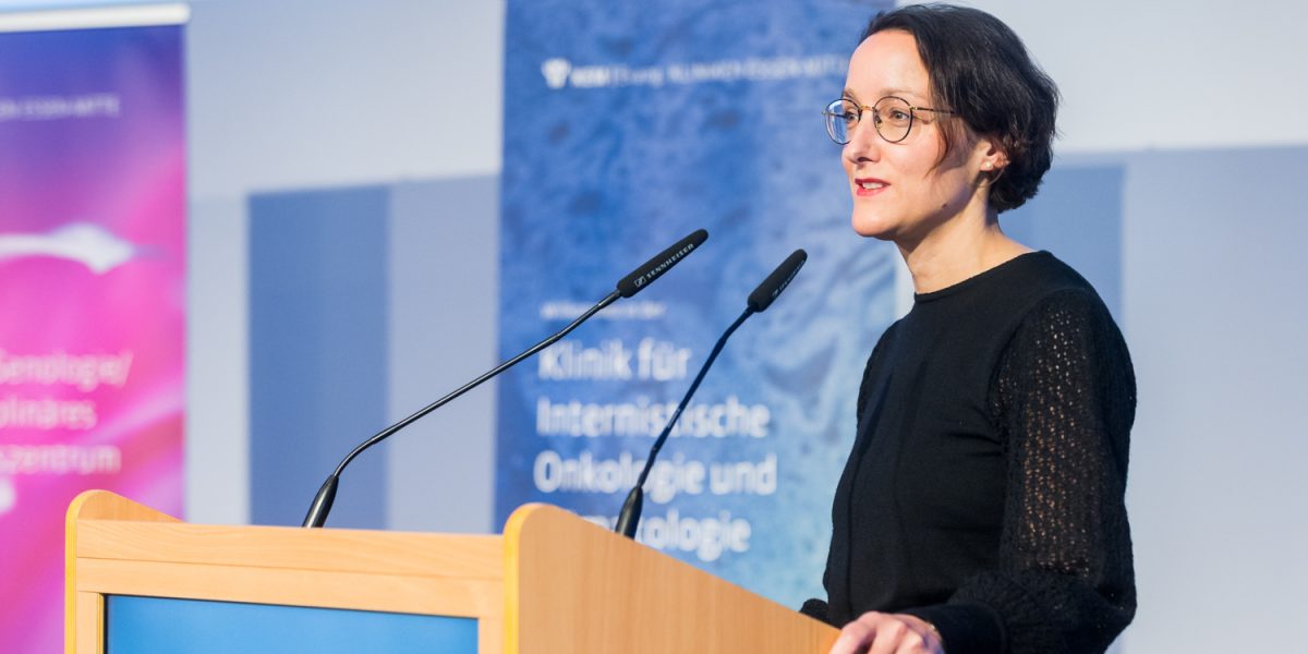 Frau hält eine Rede hinter einem Pult mit Mikrofonen. Hintergrund mit Bannern, Text auf dem Pult: 