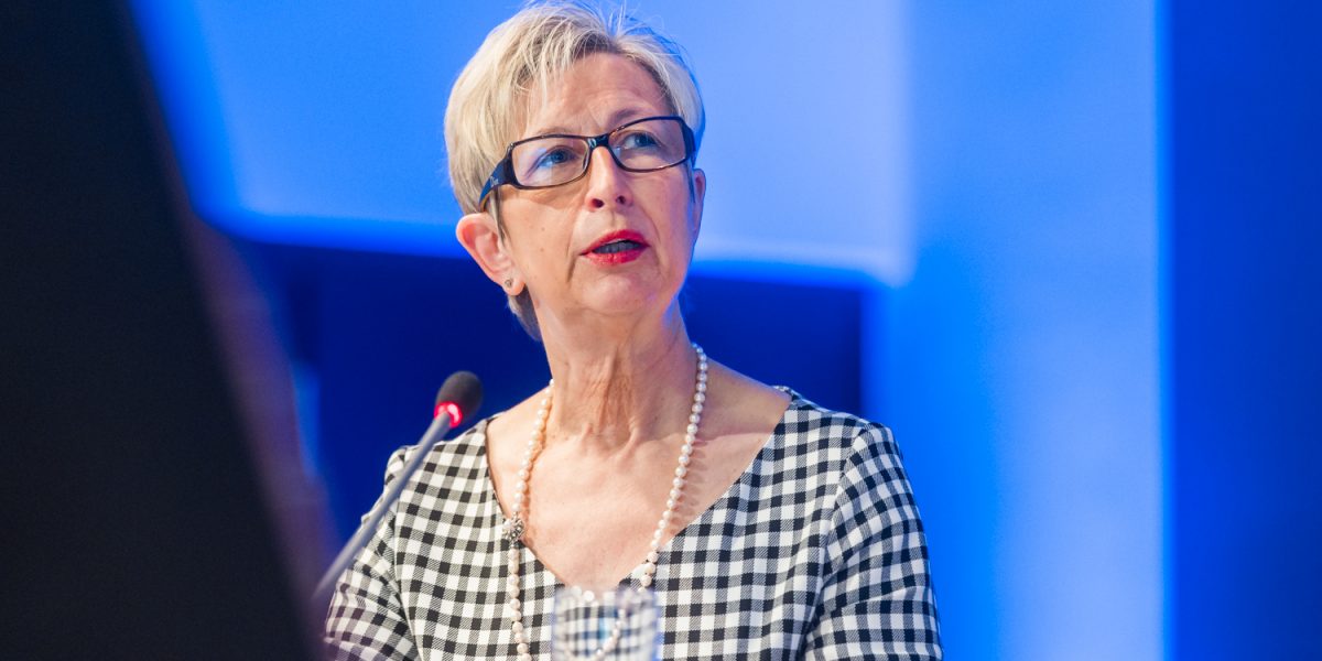 Frau mit Brille und kariertem Kleid spricht am Mikrofon vor blauem Hintergrund.