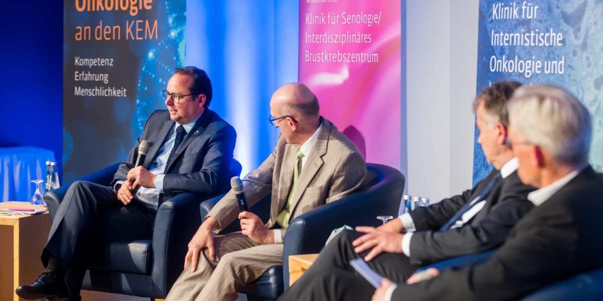 Vier Männer in Anzügen bei einer Podiumsdiskussion zur Feier von 25 Jahren Onkologie an den KEM-Kliniken.