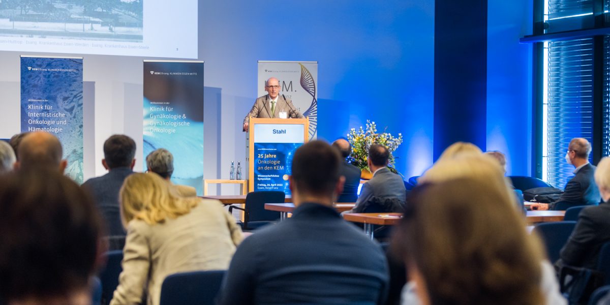 Ein Redner spricht auf einer Konferenz über Onkologie vor einem Publikum in einem blauen, beleuchteten Raum.