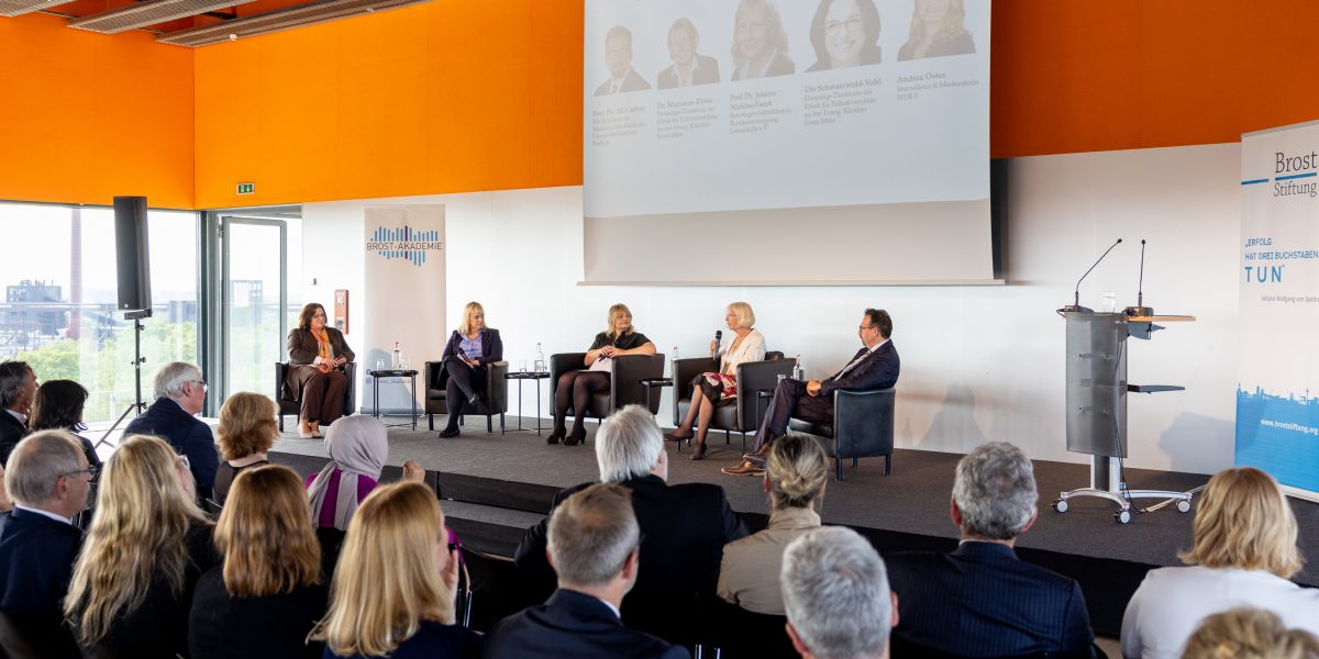 Podiumsdiskussion mit fünf Sprechern vor Publikum in einem Konferenzsaal, eine Frau hält ein Mikrofon in der Hand.