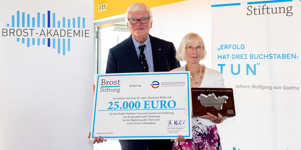 Zwei Personen halten einen Scheck über 25.000 Euro der Brost-Stiftung. Im Hintergrund sind Symbole der Brost-Akademie.