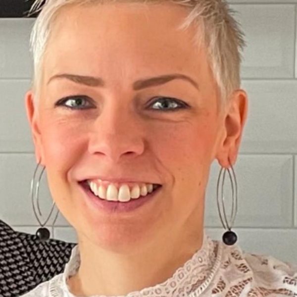 Lächelnde Person mit kurzen, blonden Haaren und Spitzenhemd vor weißer gefliester Wand.