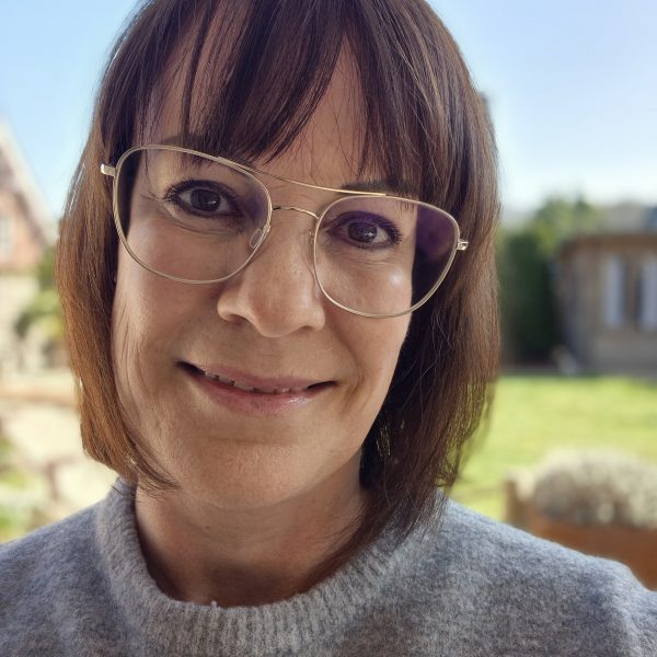 Nahaufnahme einer lächelnden Frau mit Brille und braunem Haar im Freien, blauer Himmel im Hintergrund.