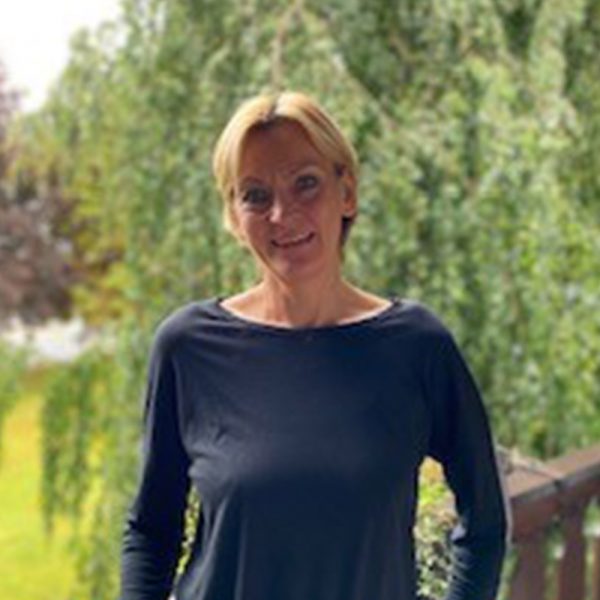 Person mit blondem Haar in schwarzem Oberteil, steht lächelnd vor einem grünen, bewaldeten Hintergrund.