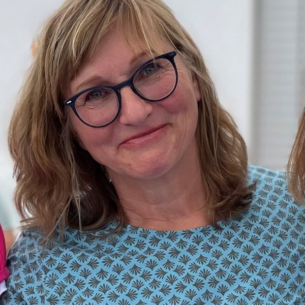 Lächelnde Frau mit Brille und schulterlangem Haar trägt ein blau-gemustertes Oberteil.