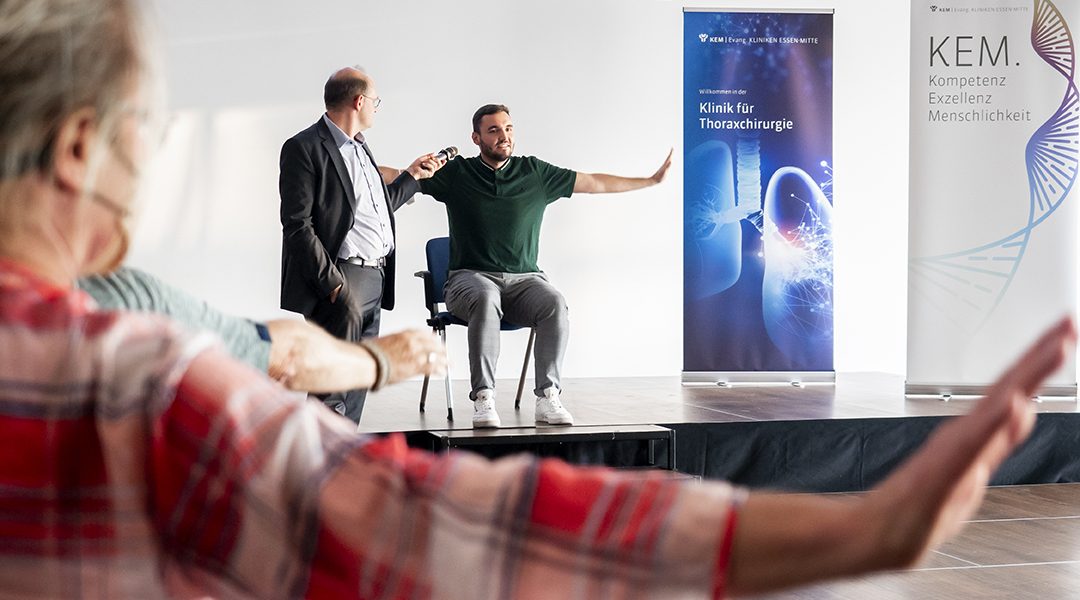 Ein Mann sitzt auf der Bühne, gestikuliert, während ein anderer ihm ein Mikrofon hält. Zuschauer bewegen die Arme.