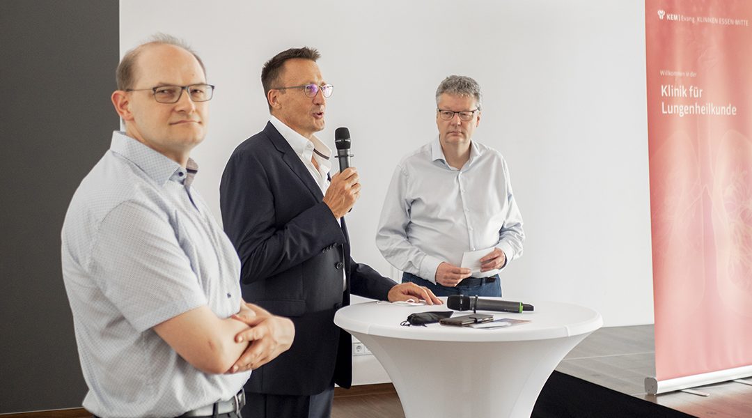 Drei Männer bei einer Präsentation am Stehtisch, einer spricht ins Mikrofon, Klinik für Lungenheilkunde im Hintergrund.