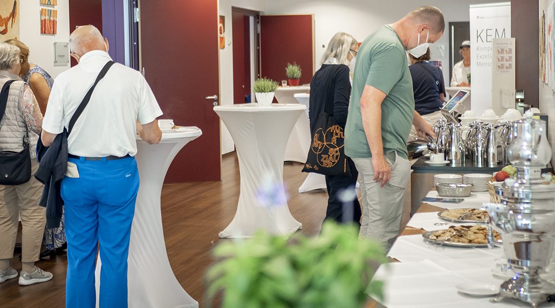 Menschen stehen an einem Buffet mit Kaffee und Snacks in einem Veranstaltungsraum mit weißen Stehtischen.