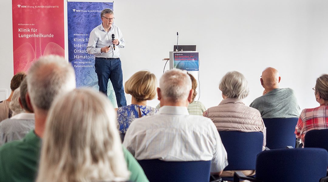 Mann hält Vortrag vor sitzendem Publikum, Hintergrund: Banner der Kliniken für Lungenheilkunde und Onkologie.