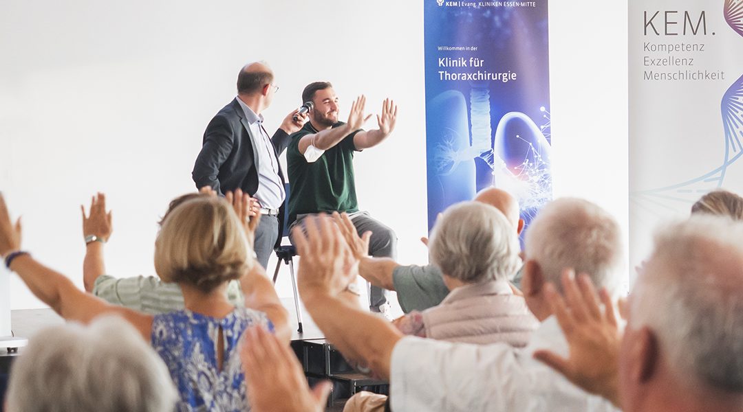 Gruppe von Menschen hebt die Arme bei einer Veranstaltung in einer Thoraxchirurgie-Klinik. Zwei Redner vorne.