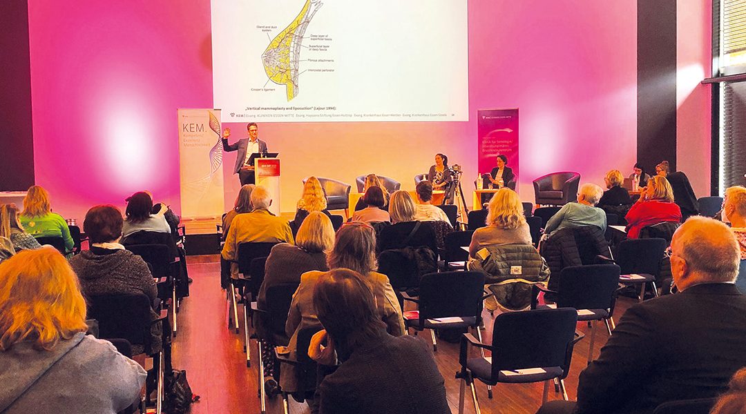Vortrag in einem Konferenzsaal mit Zuhörern, Präsentation an der Wand, Redner am Podium, pinke Wände.