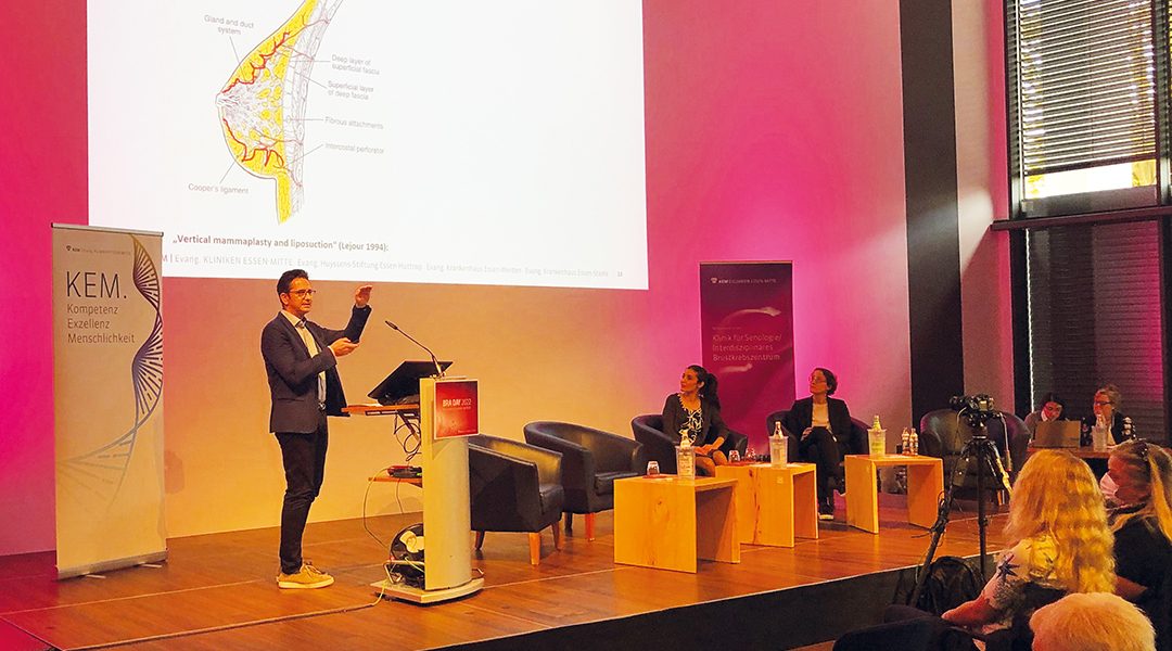 Ein Redner präsentiert eine medizinische Grafik auf einer Konferenz; Publikum und Podiumsgäste hören zu.