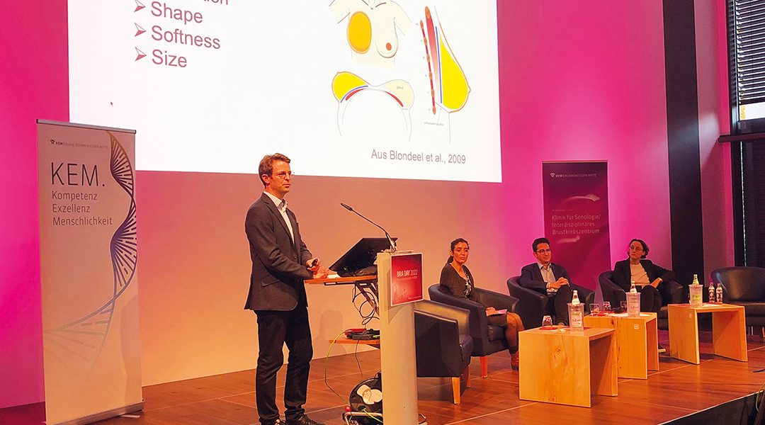 Ein Mann hält eine Präsentation über Brustrekonstruktion, während drei Personen auf einer Bühne sitzen und zuhören.