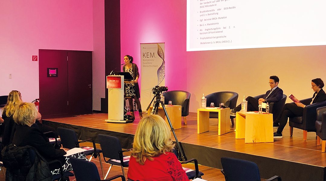 Frau hält Vortrag auf Konferenz zu Brustimplantaten, Publikum sitzt auf Stühlen, Präsentation auf Leinwand.