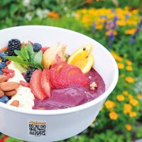 Schüssel mit Acai-Bowl, Früchten und Nüssen vor bunten Blumen im Freien.