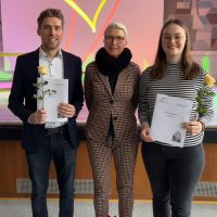 Drei Personen stehen mit Abschlussurkunden und Rosen; eine bunte Wand im Hintergrund.