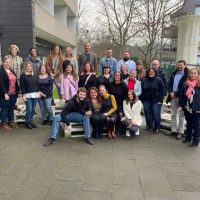 Gruppe von 24 Personen posiert draußen in legerer Kleidung vor einem Gebäude, einige sitzen auf gestapelten Paletten.