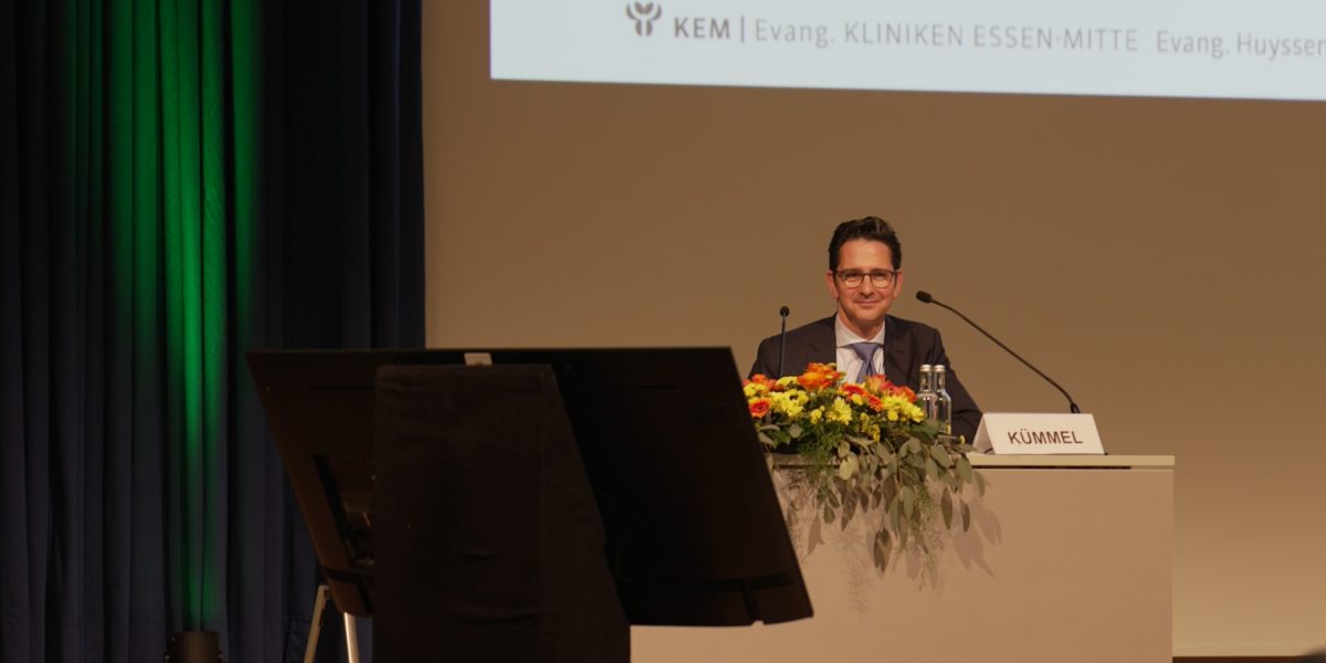 Ein Redner sitzt bei einer Konferenz auf der Bühne mit Mikrofon und Blumenarrangement, Namensschild