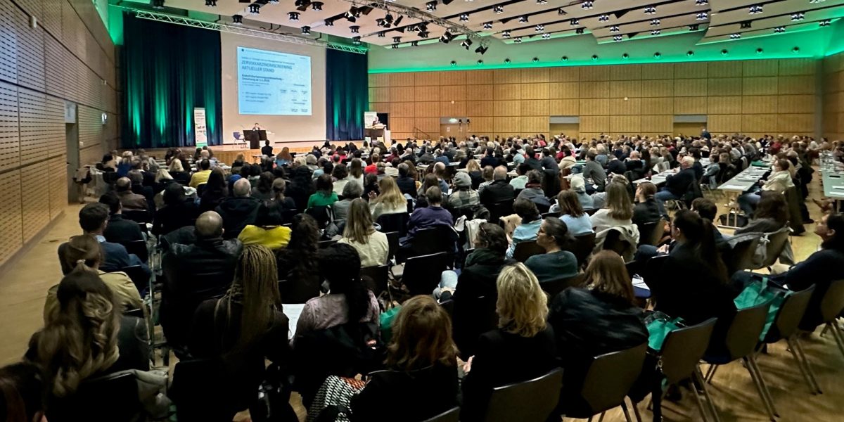 Voller Konferenzsaal, Menschen sitzen und hören einem Vortrag auf der Bühne mit großer Leinwand zu.