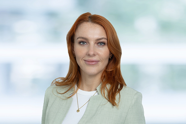 Porträt einer lächelnden Frau mit roten Haaren, hellem Blazer und weißem Oberteil vor verschwommenem Hintergrund.