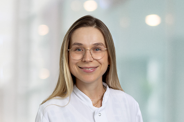 Lächelnde Frau mit Brille und weißem Kittel vor verschwommenem Hintergrund.