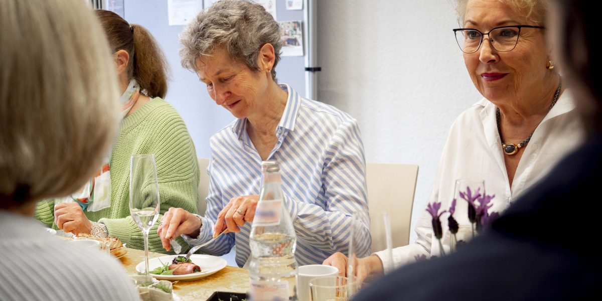 Ältere Frauen sitzen gemeinsam an einem Tisch, essen und unterhalten sich in einer hellen Umgebung.