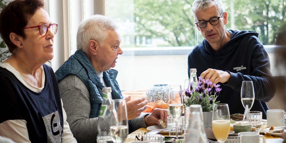 Drei Personen sitzen am Frühstückstisch, essen und sprechen miteinander, umgeben von Geschirr und Dekoration.