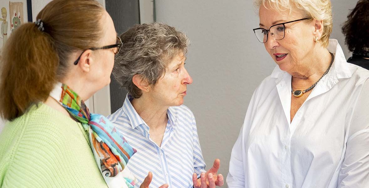 Drei Frauen führen ein Gespräch in einem Innenraum. Eine trägt ein gestreiftes Hemd, die anderen helle Blusen.