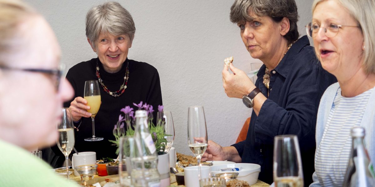 Gruppe älterer Frauen plaudert und genießt Essen und Getränke an einem gedeckten Tisch.