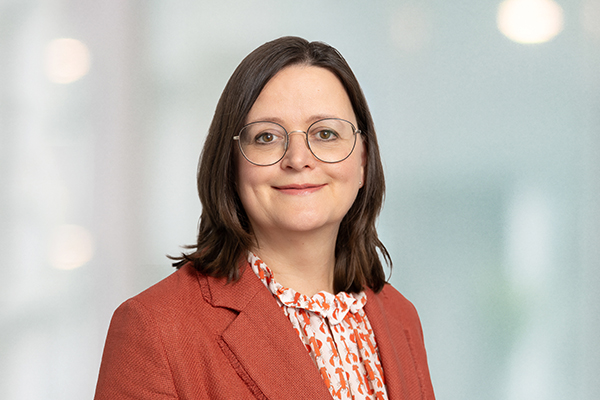 Frau mit Brille und dunklem Blazer lächelt in die Kamera vor hellem Hintergrund.