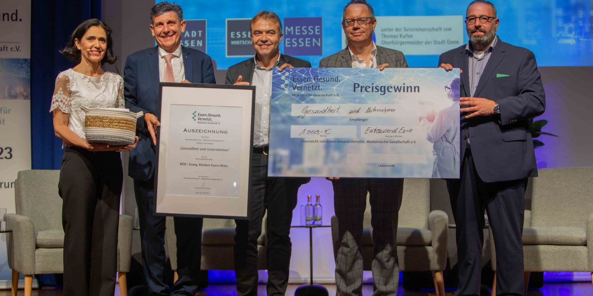 Fünf Personen stehen auf einer Bühne, halten einen großen Scheck und eine Auszeichnung in einem Rahmen.