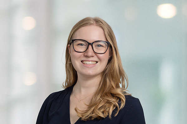 Eine lächelnde Frau mit Brille und langen Haaren vor unscharfem Hintergrund. Sie trägt ein dunkles Oberteil.