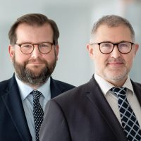 Zwei Männer in Anzügen mit Brillen posieren lächelnd vor einem unscharfen, hellen Hintergrund.