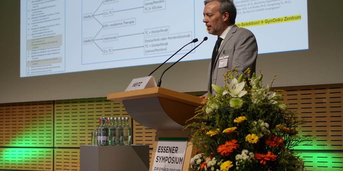 Ein Mann hält einen Vortrag beim Essener Symposium zur gynäkologischen Onkologie, neben ihm ein Blumenarrangement.
