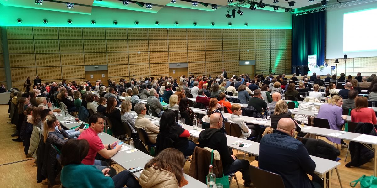 Großer Konferenzraum voller Menschen, die einem Vortrag zuhören. Die Bühne ist vorne mit grünem Licht beleuchtet.