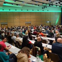 Großer Konferenzraum voller Menschen, die einem Vortrag zuhören. Die Bühne ist vorne mit grünem Licht beleuchtet.