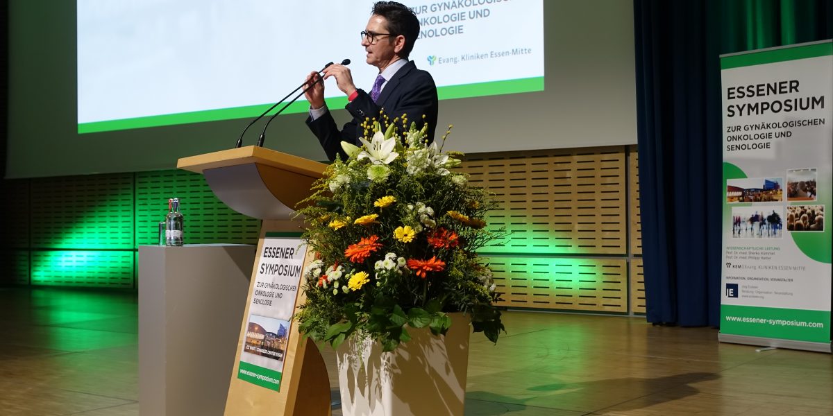 Mann spricht am Podium beim Essener Symposium, Gynäkologie-Thema; Blumenschmuck davor, Plakat im Hintergrund.